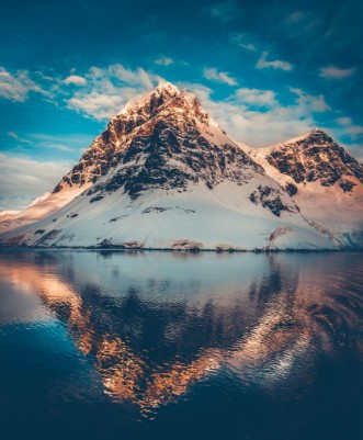 Afbeeldingen van Antarctic landscape with snow covered mountains reflected in ocean water Sunset warm light on the mountain peak blue cloudy sky in the background Beautiful nature landscape Travel background