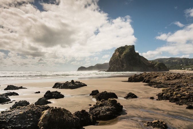 Picture of Auckland New Zealand - March 2 2017 Lion rock on Piha Beach of Tasman Sea surrounded by surf and under blue cloudy sky Forefront is sand and dispersed black volcanic rocks