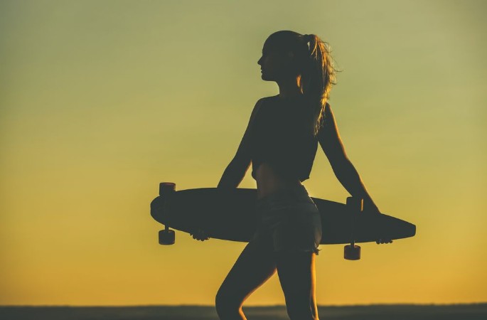 Bild på Silhouette of a beautiful sexy hipster girl in tattoos standing with a longboard against the sunset at the sea