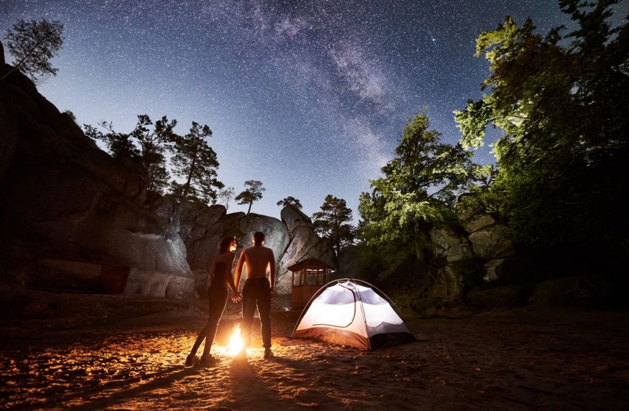 Bild på Back view silhouettes of young couple hikers man and woman having a rest beside camp bonfire and illuminated tourist tent in mountains enjoying beautiful night starry sky full of stars and Milky way