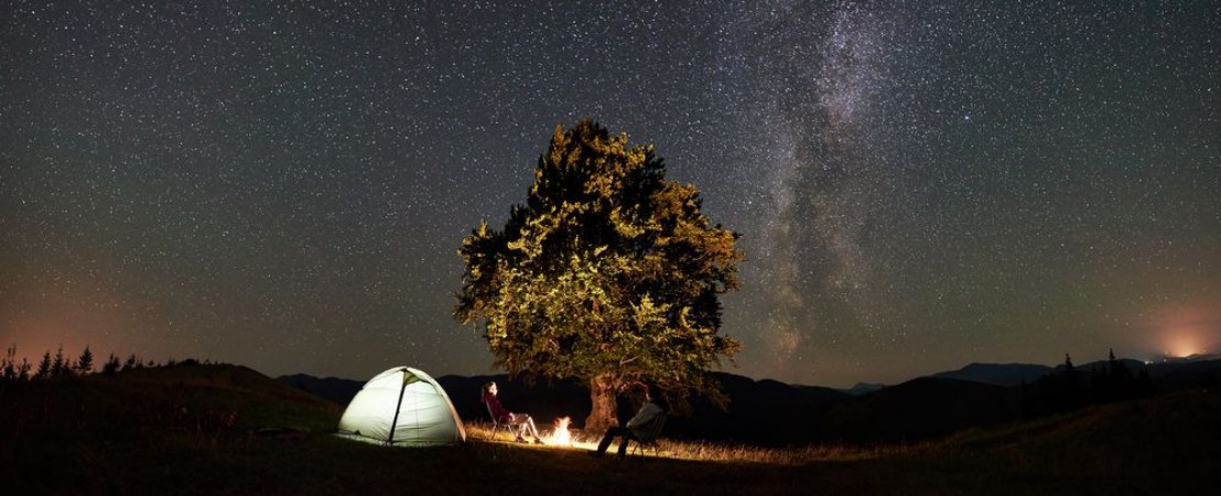Bild på Panoramic view of couple tourists resting at summer night camping in mountains Travelers sitting on chairs beside campfire illuminated tent and big tree under starry sky full of stars and Milky way